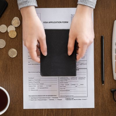 Visa application form, passport in female hands, coins, calculator, coffee on a wooden background, flat lay, visa processing, immigration.